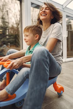 A joyful mother and son playing together on a toy car inside a sunlit room.